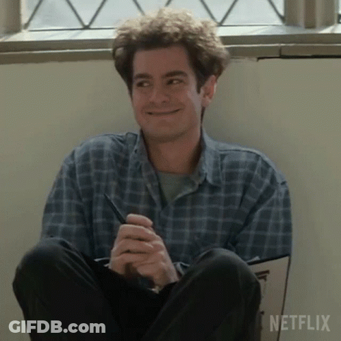 A Man In A Plaid Shirt Is Sitting On The Floor Holding A Pen And Smiling . GIF