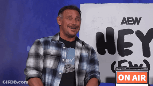 A Man In A Plaid Shirt Is Smiling In Front Of A Sign That Says Hey GIF