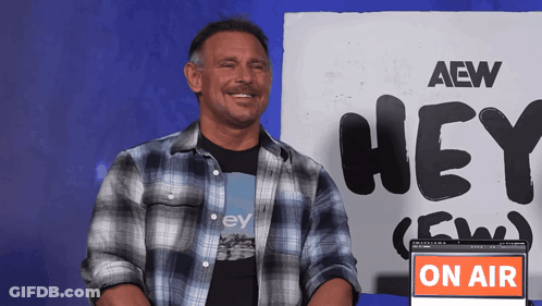 A Man In A Plaid Shirt Is Smiling In Front Of A Sign That Says Hey GIF