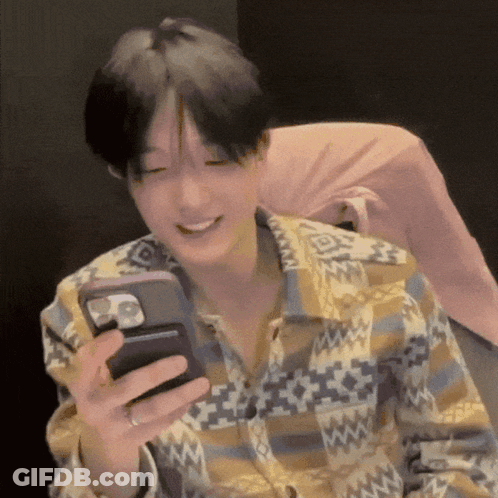 A Man In A Plaid Shirt Is Smiling While Holding A Cell Phone In His Hand . GIF