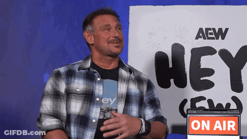 A Man In A Plaid Shirt Is Standing In Front Of A Sign That Says Hey GIF