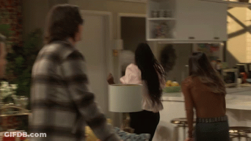 A Man In A Plaid Shirt Is Standing Next To A Woman Holding A White Lamp Shade GIF