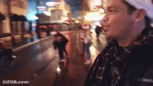 A Man In A Plaid Shirt Is Standing On A Wet City Street At Night . GIF