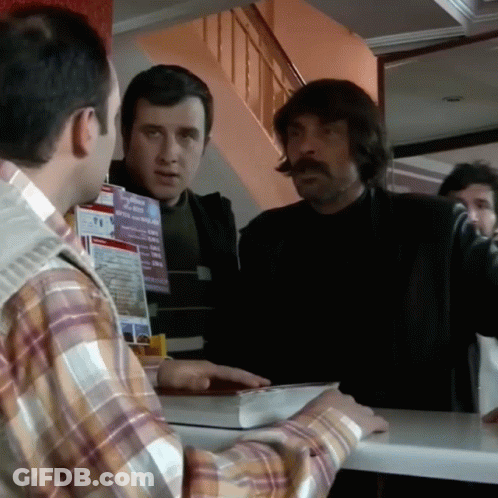A Man In A Plaid Shirt Is Talking To Another Man In Front Of A Sign That Says T.i. GIF