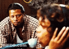 A Man In A Plaid Shirt Looks At A Woman Singing Into A Microphone GIF