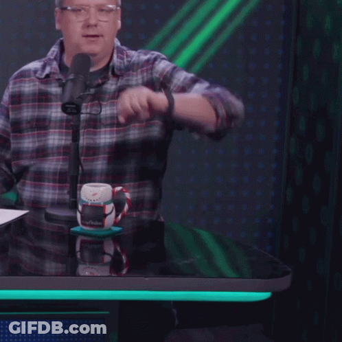 A Man In A Plaid Shirt Stands In Front Of A Microphone And Looks At His Watch GIF