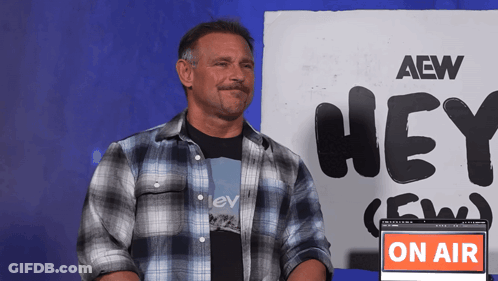 A Man In A Plaid Shirt Stands In Front Of A Sign That Says Aew Hey GIF