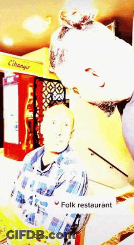 A Man In A Plaid Shirt Stands In Front Of A Sign That Says Folk Restaurant GIF