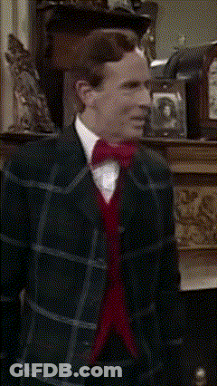 A Man In A Plaid Suit And Red Bow Tie Stands In Front Of A Fireplace GIF