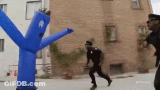 A Man In A Police Uniform Is Running Away From A Blue Blow Up Man GIF