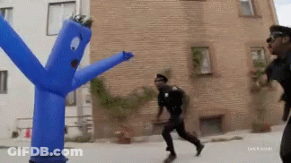 A Man In A Police Uniform Is Running Away From A Blue Inflatable Man With Arms And Legs GIF