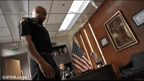 A Man In A Police Uniform Is Standing In A Room With An American Flag GIF