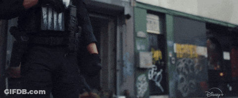 A Man In A Police Uniform Is Walking Down A Street In Front Of A Building With Graffiti On It . GIF
