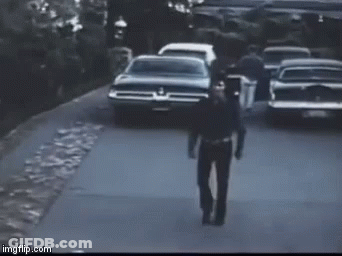 A Man In A Police Uniform Is Walking Down A Street In Front Of A Row Of Cars . GIF