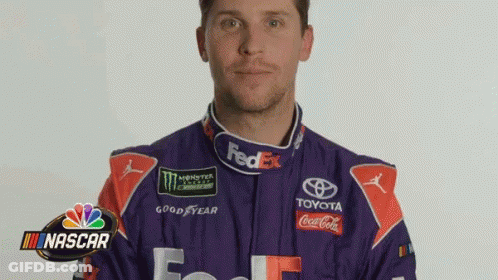 A Man In A Purple And Red Racing Suit Is Standing In Front Of A Nascar Sign . GIF