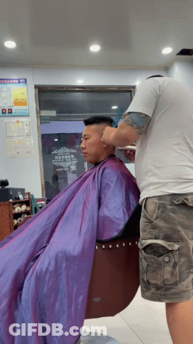 A Man In A Purple Cape Is Getting His Hair Cut By A Man In A White Shirt GIF
