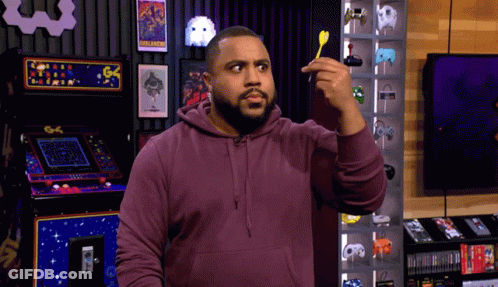 A Man In A Purple Hoodie Holds A Dart In Front Of An Arcade Machine That Says Pac Man GIF
