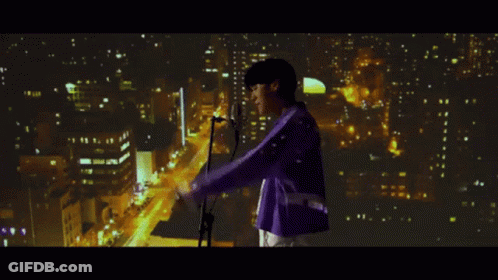 A Man In A Purple Jacket Sings Into A Microphone In Front Of A City At Night GIF