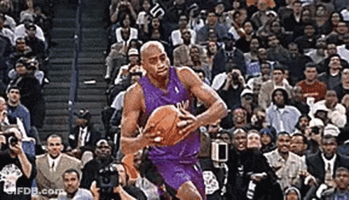 A Man In A Purple Jersey Is Holding A Basketball In Front Of A Crowd . GIF