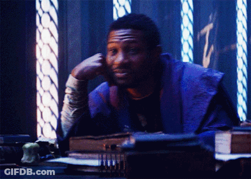 A Man In A Purple Robe Is Sitting At A Desk With His Hand On His Chin GIF