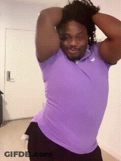 A Man In A Purple Shirt And Black Shorts Is Standing In Front Of A Door GIF