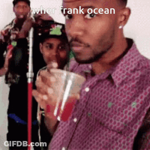 A Man In A Purple Shirt Is Drinking From A Cup While Standing In Front Of A Group Of People . GIF