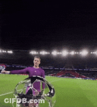 A Man In A Purple Shirt Is Holding A Soccer Ball On A Field At Night GIF