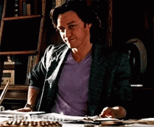 A Man In A Purple Shirt Is Sitting At A Desk With A Bookshelf In The Background GIF