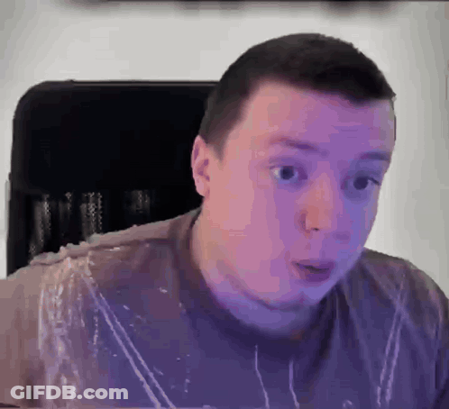 A Man In A Purple Shirt Is Sitting In A Chair With A Surprised Look On His Face . GIF