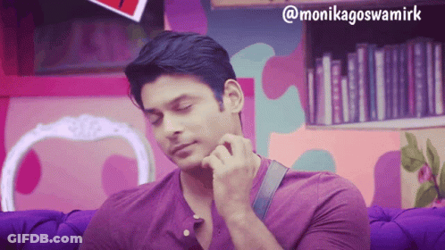 A Man In A Purple Shirt Is Sitting On A Purple Couch In Front Of A Bookshelf GIF
