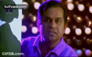 A Man In A Purple Shirt Is Standing In Front Of A Blurry Background Of Lights . GIF