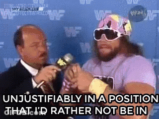 A Man In A Purple Shirt Is Talking Into A Microphone While A Man In A Suit Looks On . GIF