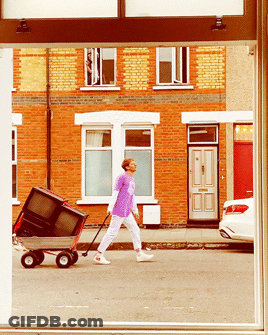 A Man In A Purple Shirt Pushes A Wagon With A Television On It GIF
