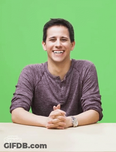 A Man In A Purple Shirt Sits At A Table With His Hands Folded In Front Of A Green Screen GIF