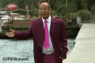 A Man In A Purple Suit And Pink Tie Is Standing In Front Of A Trt Sign GIF