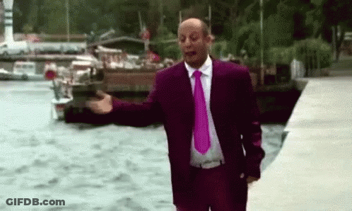 A Man In A Purple Suit And Pink Tie Is Standing Next To A Body Of Water With His Arms Outstretched . GIF