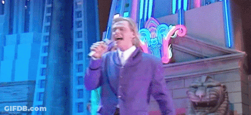 A Man In A Purple Suit Is Singing Into A Microphone On A Stage GIF