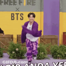 A Man In A Purple Sweater And Purple Pants Is Dancing In Front Of A Free Fire Sign GIF