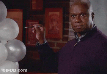 A Man In A Purple Sweater Is Standing In Front Of Balloons And Holding A Pen In His Hand . GIF