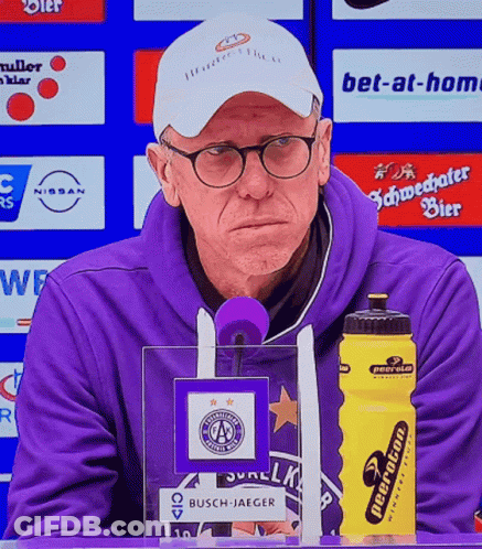 A Man In A Purple Sweatshirt Stands In Front Of A Microphone Next To A Yellow Water Bottle That Says Neeraton GIF