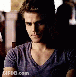 A Man In A Purple T-Shirt Is Sitting In A Dark Room . GIF