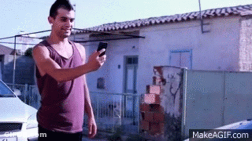 A Man In A Purple Tank Top Is Holding A Cell Phone In Front Of A White Car . GIF