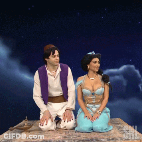A Man In A Purple Vest And A Woman In A Blue Dress Are Sitting On A Carpet That Says Saturday Night Live GIF