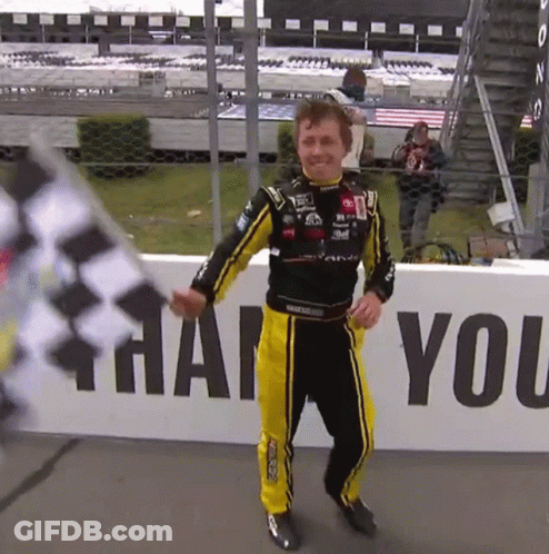 A Man In A Race Suit Holds A Flag In Front Of A Sign That Says Thank You GIF