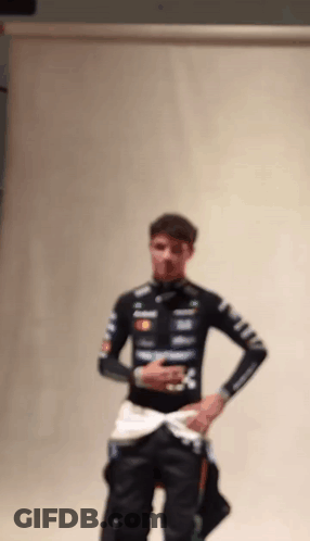 A Man In A Racing Suit Is Standing In Front Of A White Wall With His Hands In His Pockets . GIF