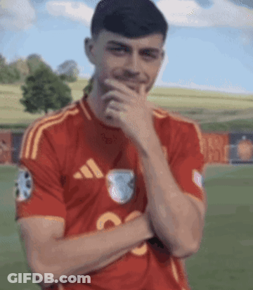 A Man In A Red Adidas Shirt Is Covering His Mouth With His Hand While Standing On A Field . GIF