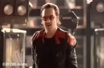 A Man In A Red And Black Jacket And Sunglasses Is Standing In A Room . GIF