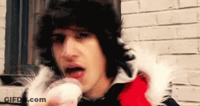 A Man In A Red And Black Jacket Is Eating A Cupcake In Front Of A Brick Wall . GIF