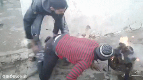 A Man In A Red And Black Striped Shirt Is Kneeling Down Next To A Fire . GIF