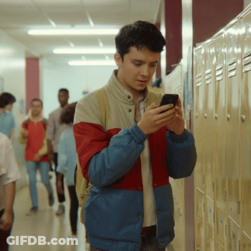 A Man In A Red And Blue Jacket Looks At His Phone In A Hallway GIF